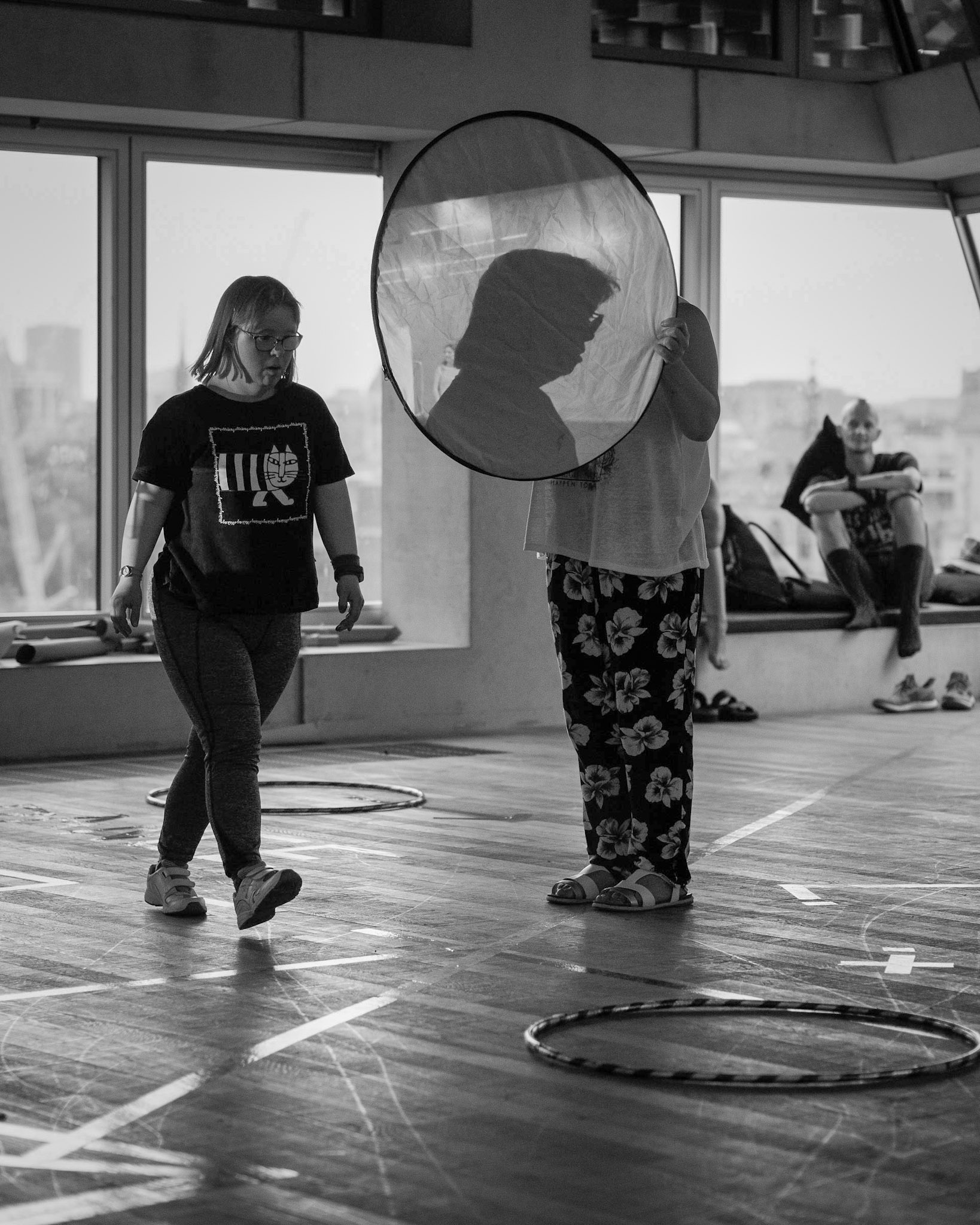Black & White photo of Bethan Kendrick walking past Jackie Ryan holding a hoop with paper stretched across it with the shadow of Bethan on it